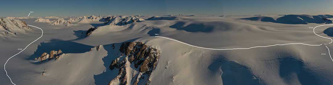 A drone panorama showing part of the Inglefield Mountains of Ellesmere Island. At the far left is Peak 1 (1,450m, 77°47.669'N, 79°14.991'W). The white line shows the 2019 party’s ski path (toward the camera), dips out of the picture, and then comes up on the far right to reach Camp 10. From Camp 10 the team ascended Peak 3 (1,410m, 78°2.722'N, 80°4.599' W, (center of image). The ski route away from Camp 10 is visible on the far right. 