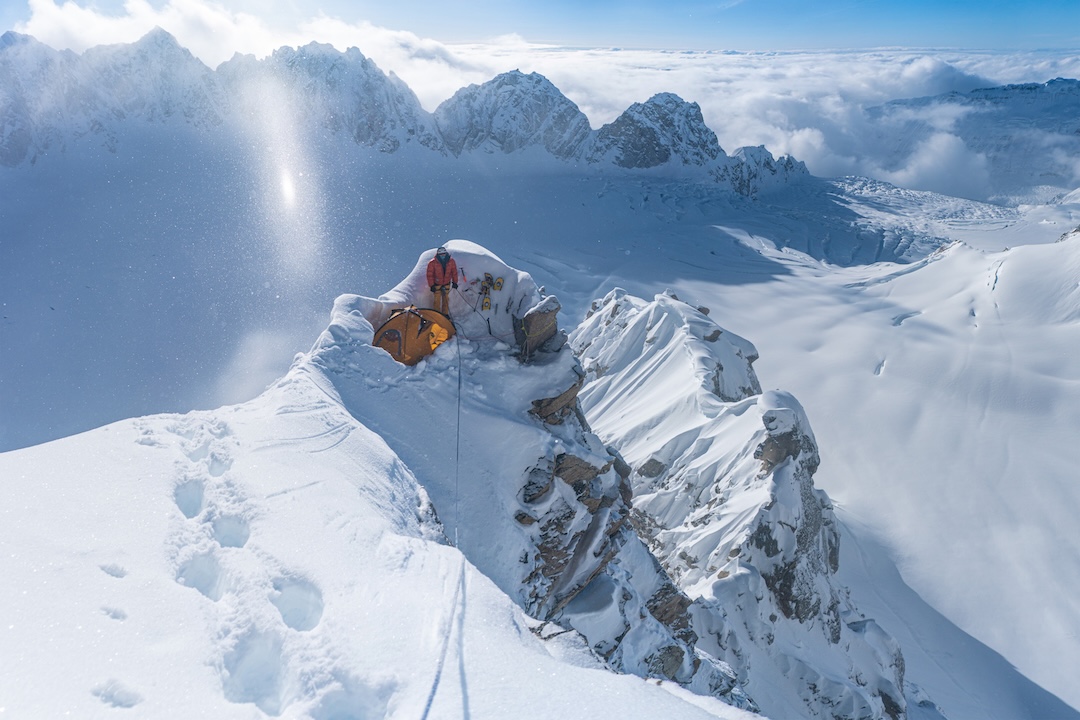 Chris Dewey at a bivy on the Southeast Spur of Begguya. One pitch above, Ben and Chris Dewey abandoned their attempt on the spur. The cirque below the Southeast Spur, where the brothers were dropped by Talkeetna Air Taxi, is in the background in shadow. The summit of the Water Tower can barely be seen at the far left of the ridge behind. 