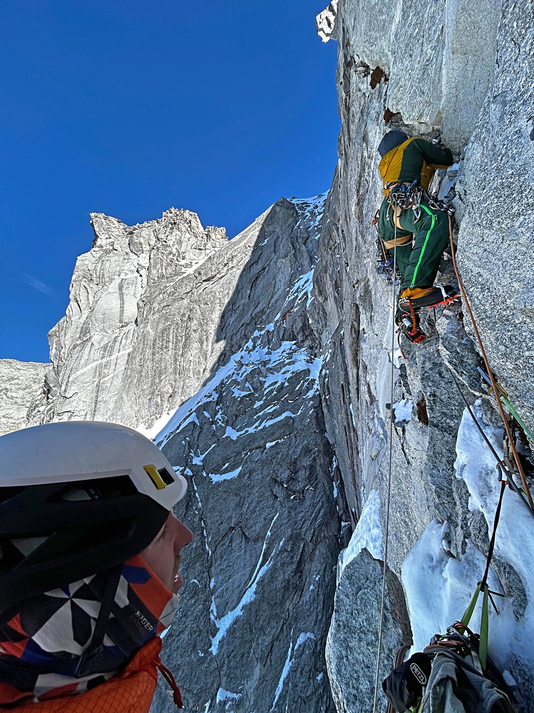 Watched by Filippo Sala, Silvan Schüpbach starts up a hard mixed crack system in the lower half of the north face of Punta Pioda di Sciora. Above, in sunlight, is the summit beak of the Pioda, while to the left is the west wall of the Sciora Dafora, with three aid and free routes (VI A3) from the 60s and 70s.