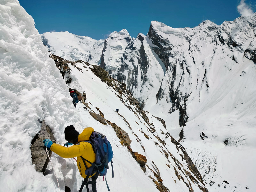 Moving along fixed ropes on the traverse in the middle of the 2023 Indian ascent of the west face of Meru. Behind lies part of the ridge connecting Bhartekhuta (6,578m, distant snowy peak on left) to Thalay Sagar. 